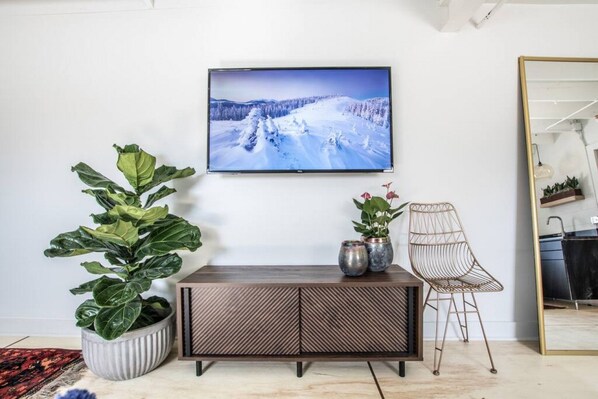 Una sala de estar con un televisor grande en la pared, un mueble de madera, una planta grande en maceta, una silla y un espejo.