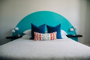 Ein Bett mit einem petrolfarbenen Kopfteil, zwei weißen Kissen und einer bunten Tagesdecke.