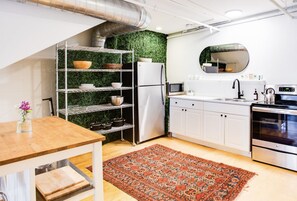 Eine moderne Küche mit weißen Schränken, einem Edelstahl-Kühlschrank, einer Mikrowelle und einem Backofen. Eine grüne Wand mit einem Regal, das Schüsseln und Körbe beherbergt. Ein Holztisch mit einer Blumenvase steht im Vordergrund.