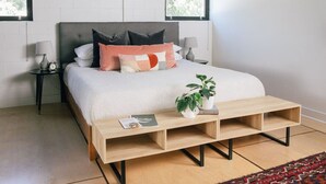 Um quarto com uma cama, mesas de cabeceira, um criado-mudo, uma mesa de centro de madeira e uma planta em vaso.