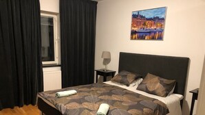 Ein ordentlich bezogenes Bett mit Bettwäsche mit Blattmuster, ein Fenster mit dunklen Vorhängen und ein Stadtbild an der Wand.