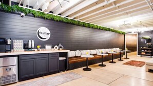 Un área de descanso moderna con pared de madera oscura, un cartel de Stellar Suites, una estación de café y un espacio de estar con mesas redondas y bancos.