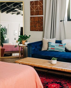 A cozy room with a blue sofa, a wooden coffee table, and a large mirror. There are plants on the table and a red patterned rug on the floor.