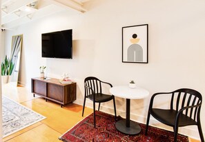 Un salon moderne avec un téléviseur à écran plat, deux fauteuils noirs, une table ronde, un tapis rouge à motifs et une œuvre d’art minimaliste accrochée au mur.