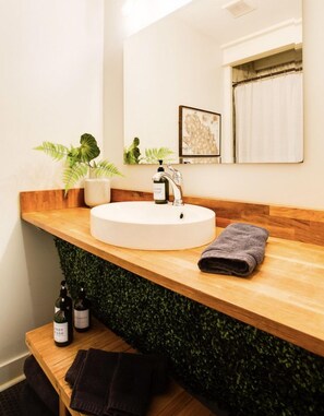 Une salle de bain avec un lavabo rond blanc, un plan de toilette en bois, un miroir et un mur vert. On y trouve deux flacons de gel douche et une serviette sur le plan de toilette.