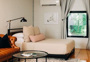A modern bedroom with a bed, a leather sofa, a round coffee table, and a window with a view of greenery.