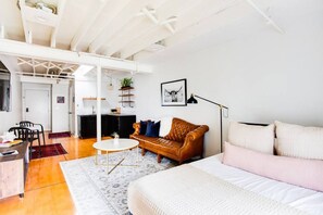 Un salon moderne avec un canapé en cuir marron, un divan blanc et une table basse ronde. On aperçoit à l’arrière-plan un coin cuisine avec un plan de travail noir et des placards blancs.