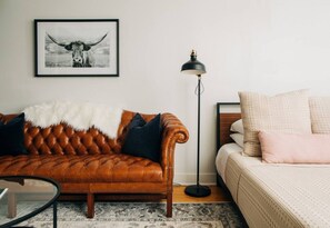 A living room with a brown leather sofa, a floor lamp, and a bed with a white comforter.