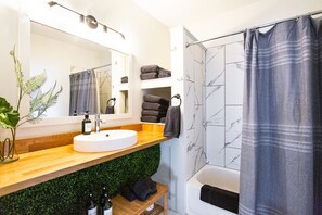 A modern bathroom with a wooden vanity, a round sink, a mirror, a shower with a marble tile wall, and a grey shower curtain.
