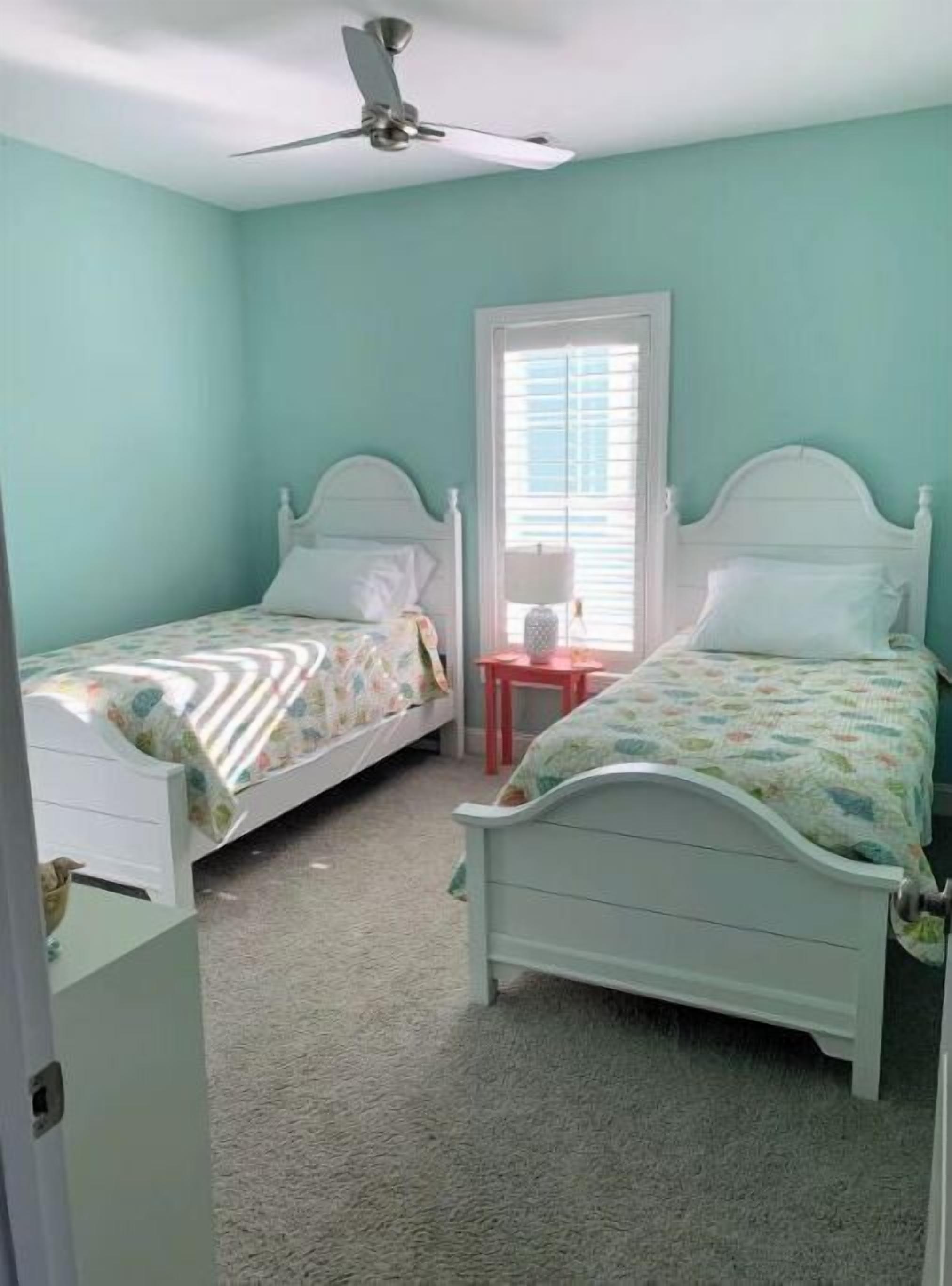 A bedroom with two single beds, a ceiling fan, a window with blinds, and a small red table.