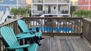 Eine Holzterrasse mit türkisfarbenen Adirondack-Stühlen, einem Pool und einem mehrstöckigen Gebäude im Hintergrund.