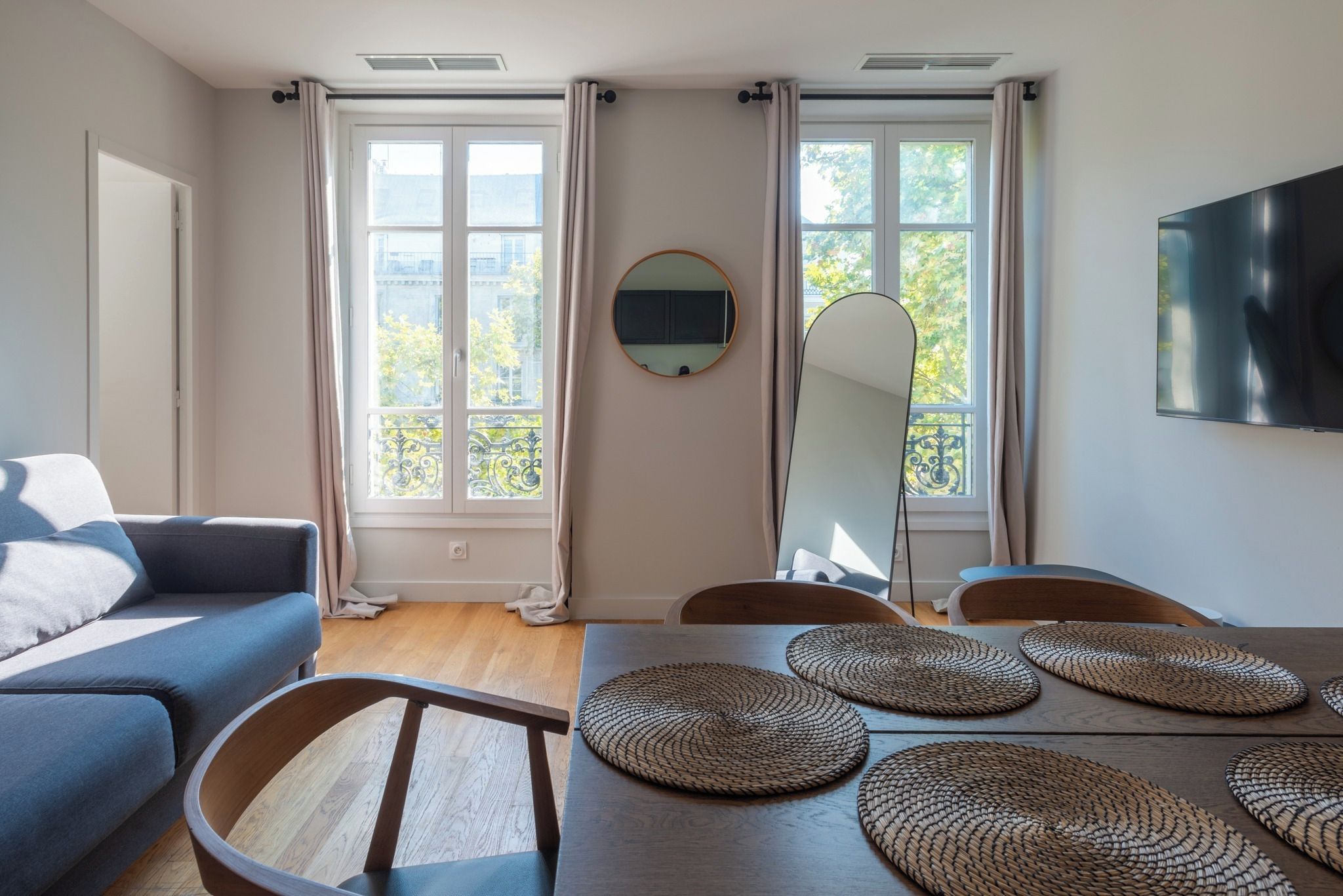 Un salon moderne avec un canapé, une table à manger et des chaises. Il y a deux fenêtres avec des rideaux, un téléviseur à écran plat fixé au mur et un miroir rond au-dessus de la table.