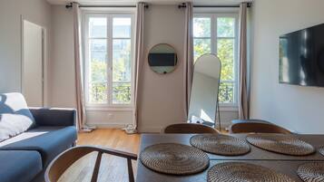 Un salon moderne avec un canapé, une table à manger et des chaises. Il y a deux fenêtres avec des rideaux, un téléviseur à écran plat fixé au mur et un miroir rond au-dessus de la table.
