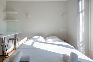 A minimalist hotel room with a bed, two bedside tables, a desk, and a window.