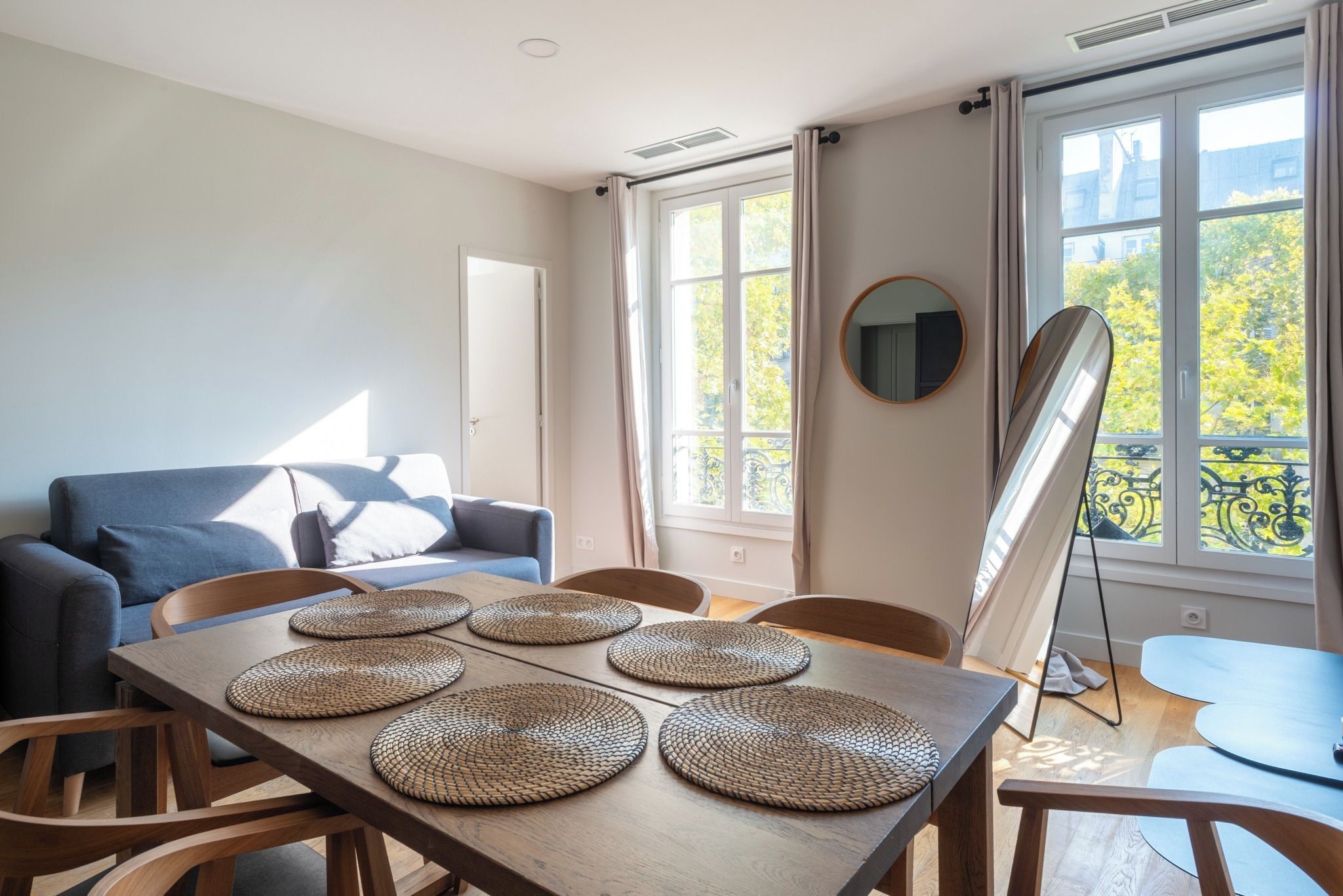 Un salon moderne avec un canapé, une table à manger avec des sets de table et de grandes fenêtres avec des rideaux.