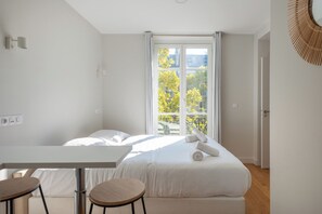 A modern hotel room with a bed, a table, two chairs, a window with a view of trees, and a door. - Bright and Stylish Paris Apartment (Paris)