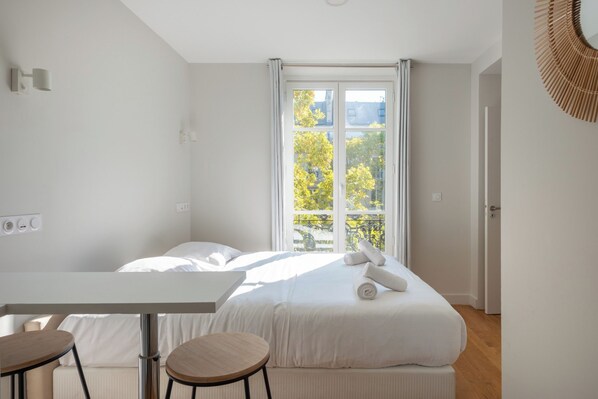 A modern hotel room with a bed, a table, two chairs, a window with a view of trees, and a door.