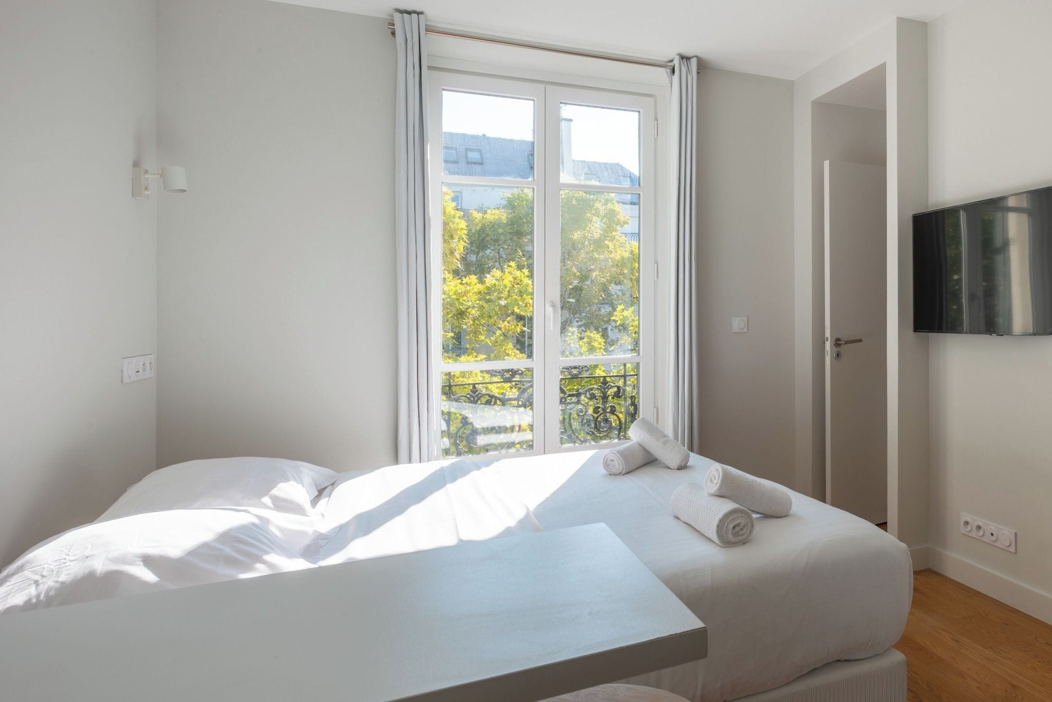 Une chambre à coucher avec un lit, une fenêtre avec des rideaux, un téléviseur à écran plat fixé au mur et un plancher en bois.