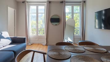 Un salon moderne avec un canapé, une table à manger et des chaises. Il y a deux fenêtres avec des rideaux, un téléviseur à écran plat fixé au mur et un miroir rond au-dessus de la table.