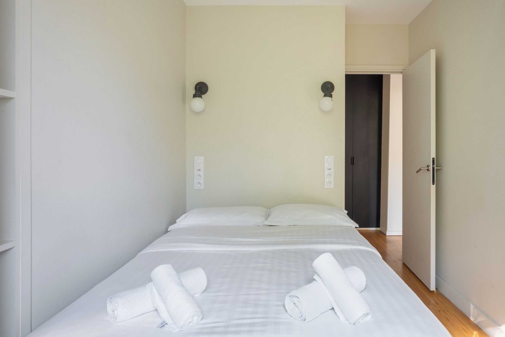 A single-bed room with a door to the right, a light switch, and a power outlet on the wall.