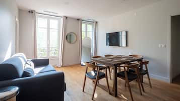 Ein modernes Wohnzimmer mit einer Couch, einem Esstisch mit Stühlen, einem flachbildfernseher an der Wand und großen Fenstern mit Vorhängen.