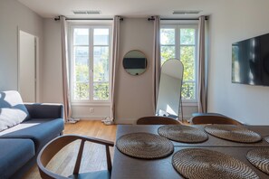 A modern living room with a sofa, dining table, and chairs. There are two windows with curtains, a flat-screen TV mounted on the wall, and a round mirror above the table.
