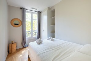 A bedroom with a bed, a bedside table, a wardrobe, a window with curtains, and a wall-mounted mirror.