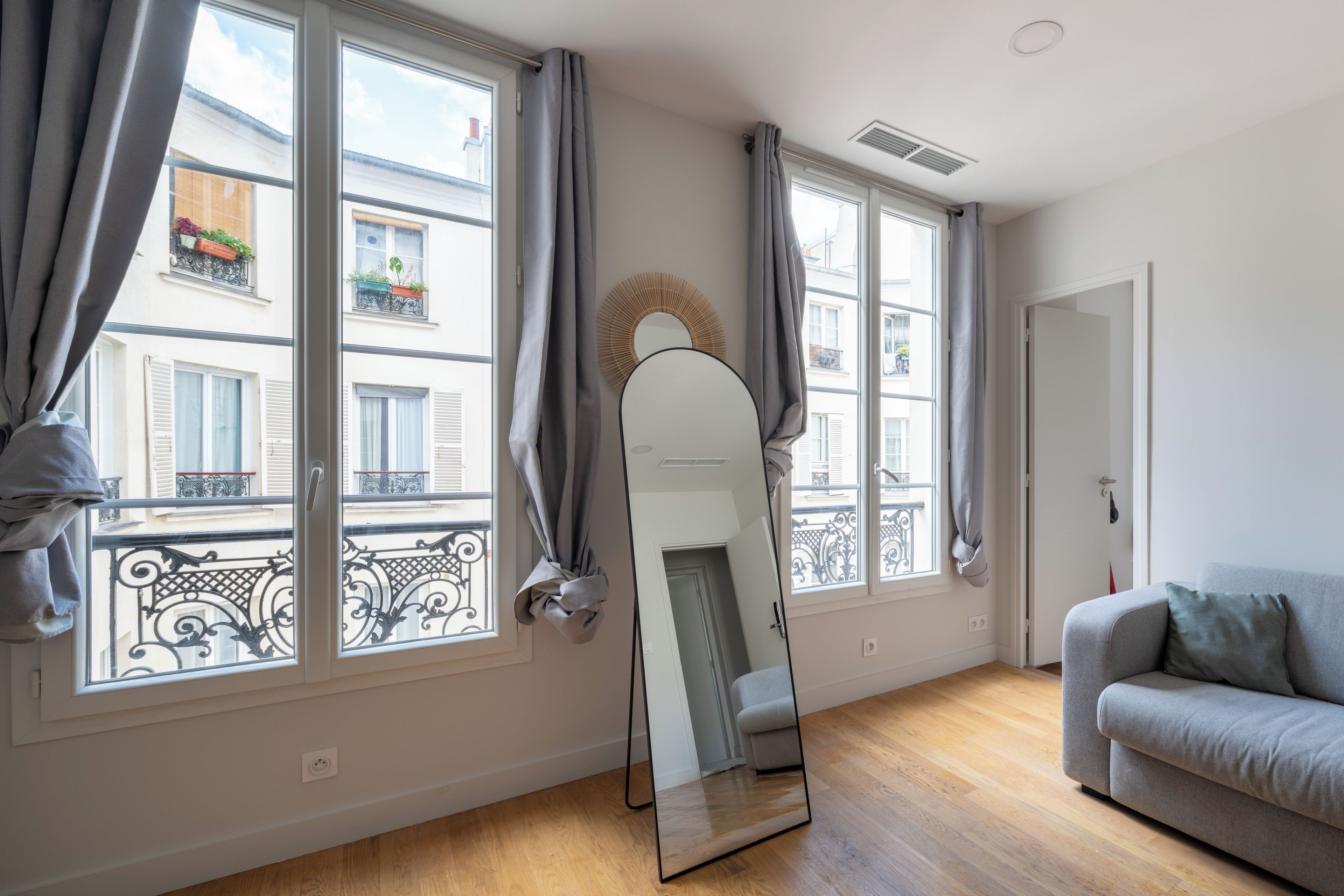 Une pièce avec un canapé gris, un grand miroir et deux fenêtres avec des rideaux gris.