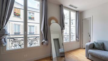 Une pièce avec un canapé gris, un grand miroir et deux fenêtres avec des rideaux gris.