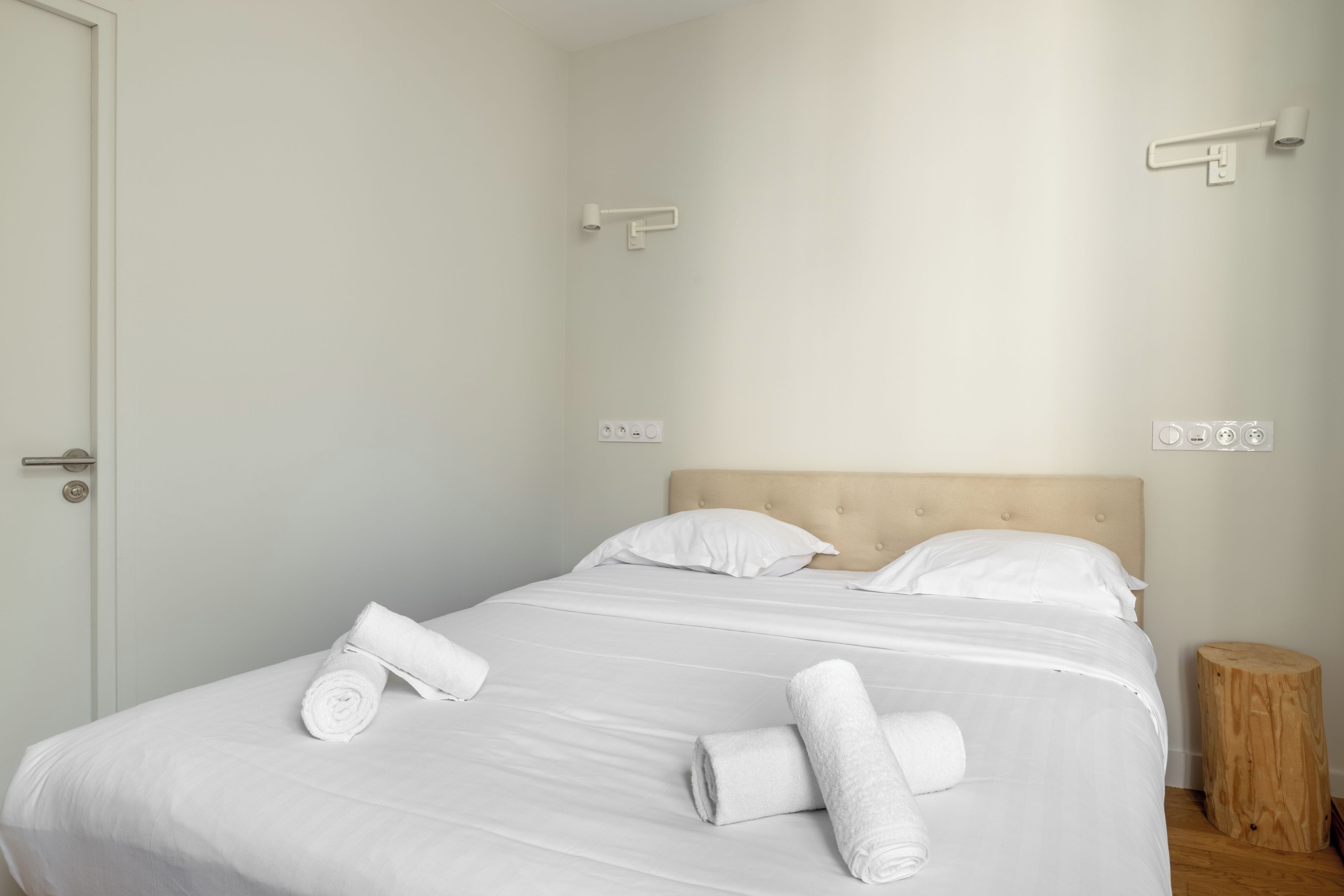 Un lit bien fait, avec du linge de lit blanc et des serviettes pliées, une tête de lit en bois et un petit tabouret en bois placé à côté du lit.