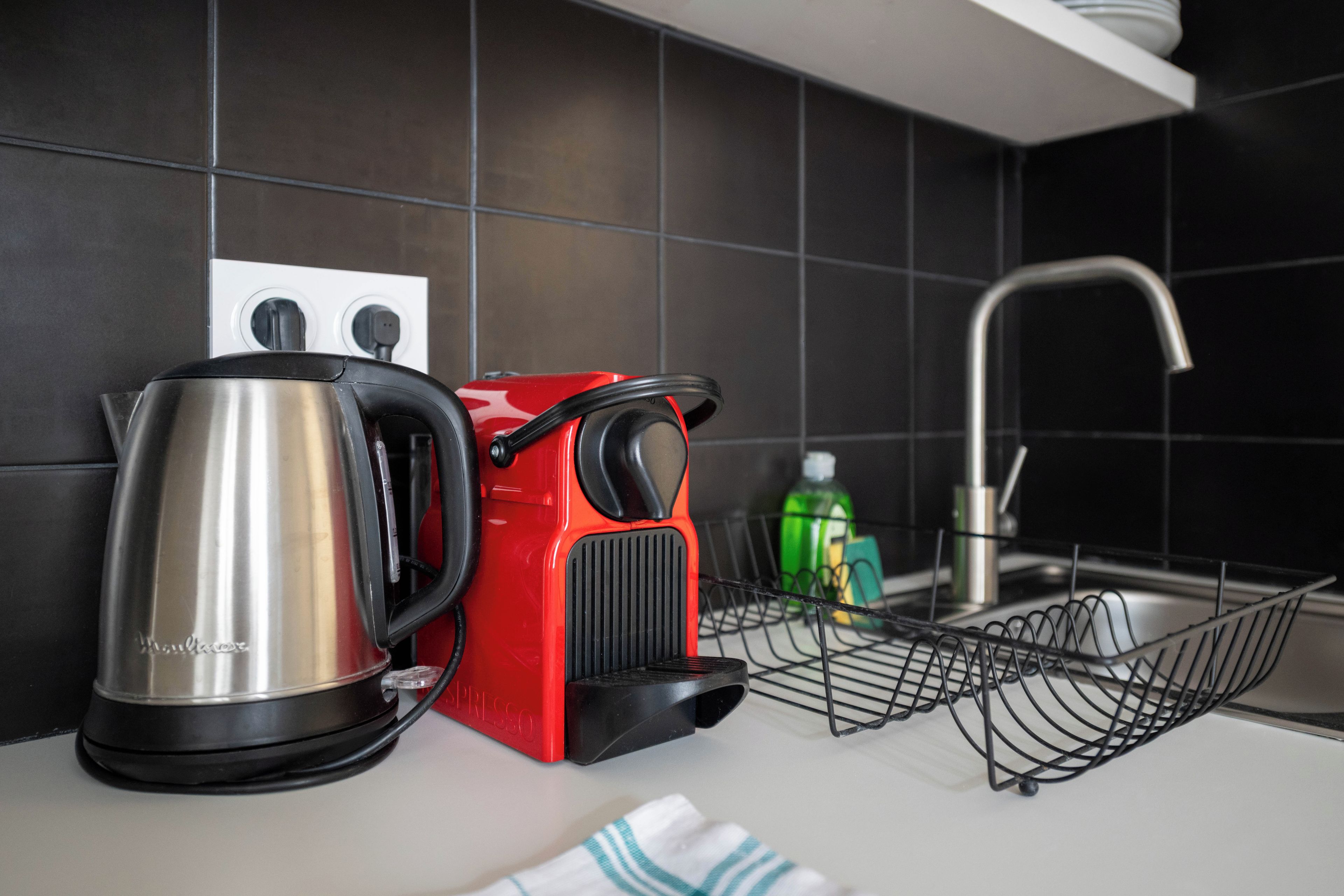 Een keuken met een waterkoker, een rode koffiemachine, een groene fles en een metalen rek.
