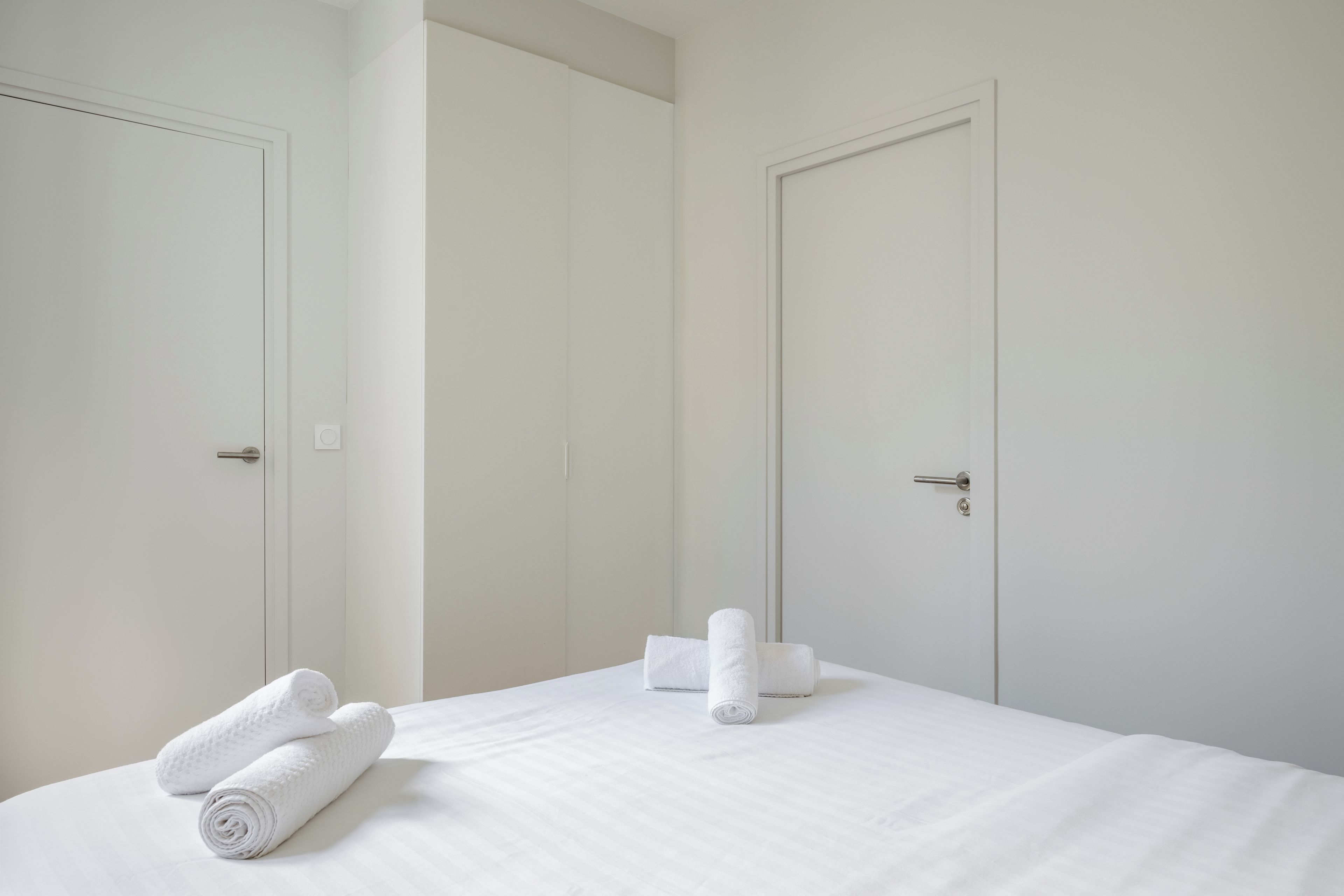 A hotel room with a bed, two doors, and two rolled towels on the bed.