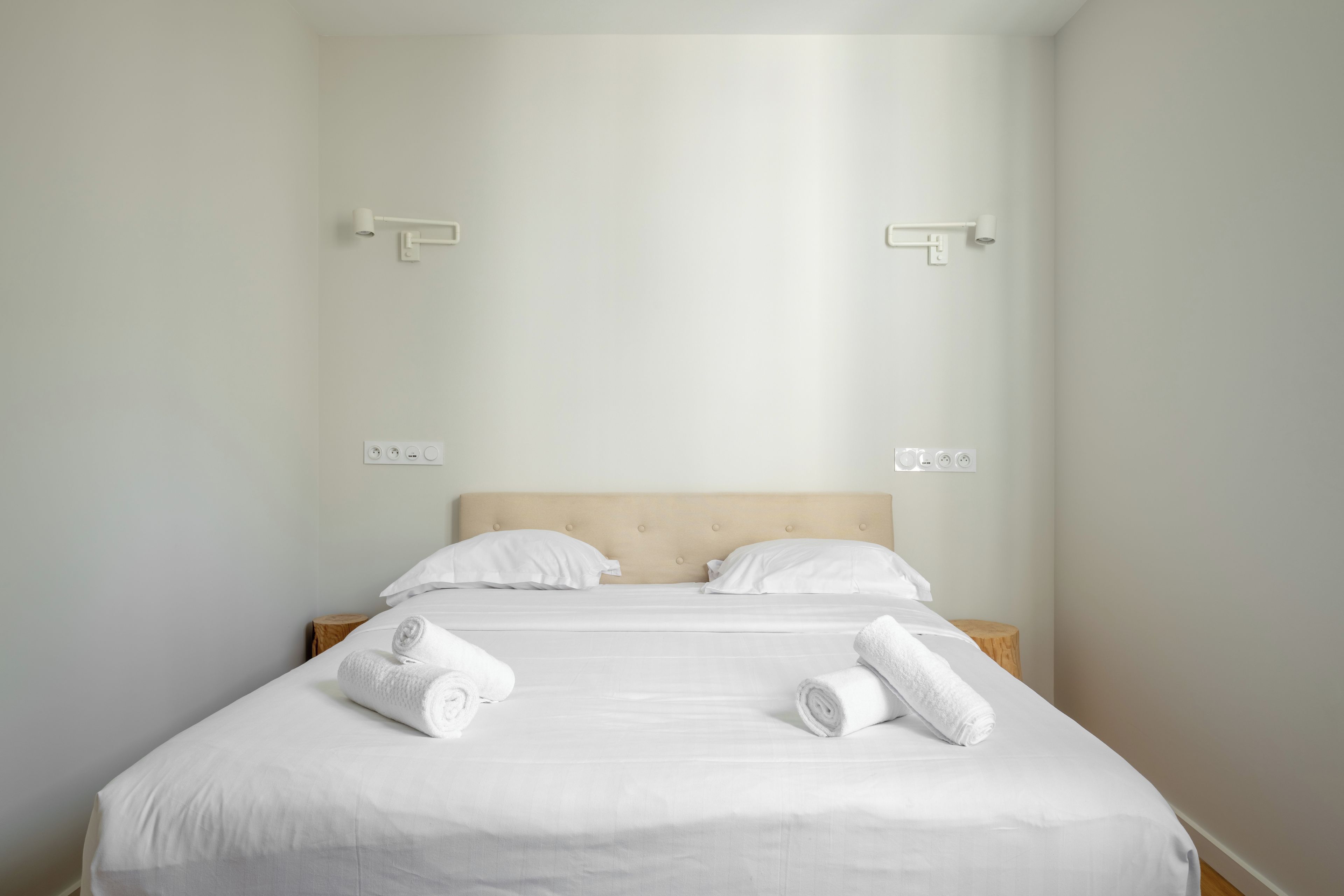 Un lit bien fait, avec du linge de lit blanc et deux serviettes enroulées posées sur le côté, une table de chevet en bois et un luminaire fixé au mur.
