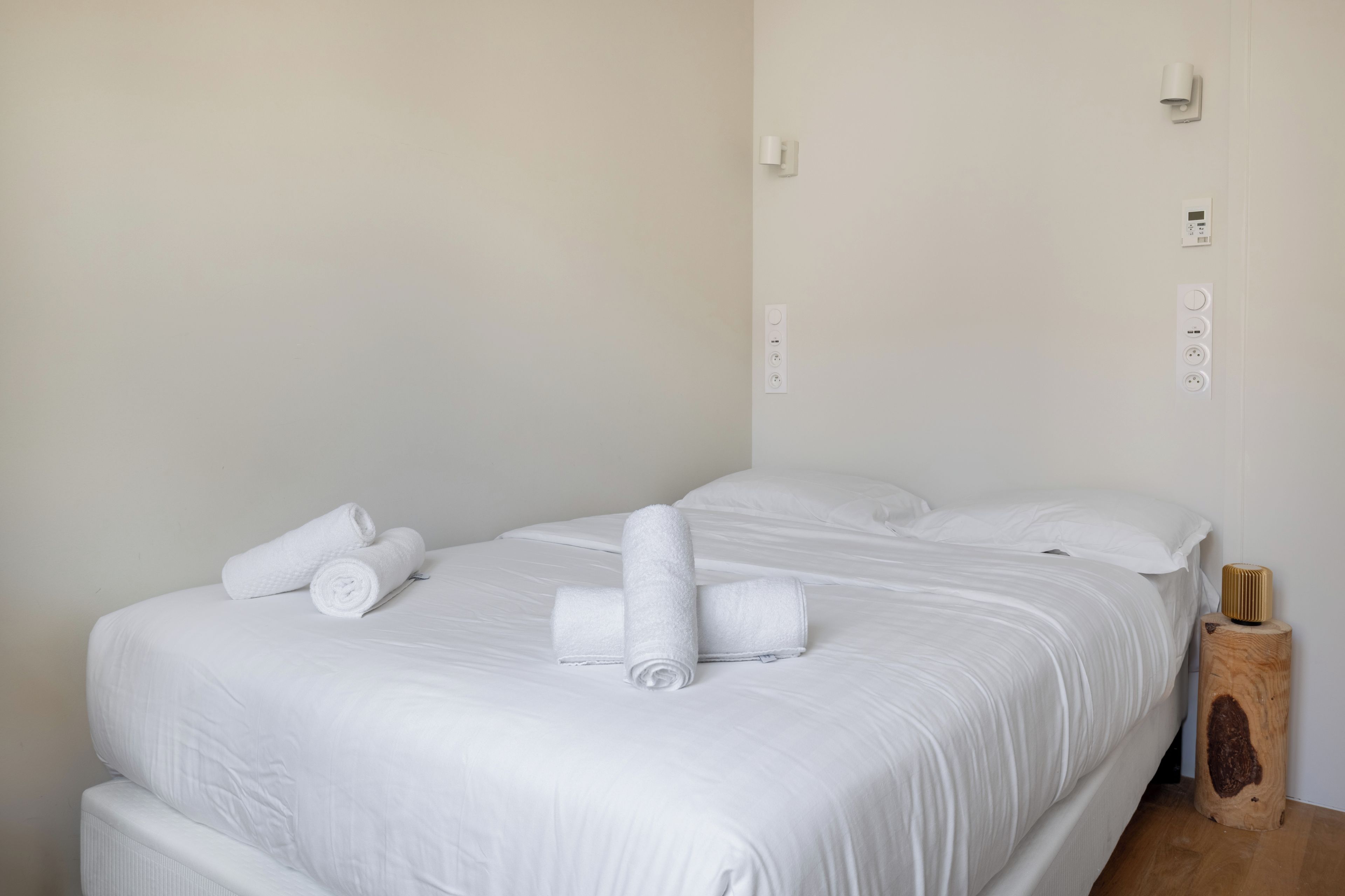 Ein Hotelzimmer mit einem Bett, zwei aufgerollten Handtüchern, einem Nachttisch aus Holz mit einem Lautsprecher und einer Steckdose.