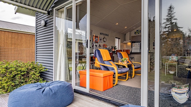 A modern outdoor seating area with blue bean bags and orange chairs, a wooden fence, and a glass door leading to an interior space with a kitchen and dining area. - Alpine Way Cottage - Ohakune Holiday Home (Ohakune)