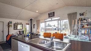 A kitchen with a large island, a dishwasher, and a dining area with chairs and a table. - Alpine Way Cottage - Ohakune Holiday Home (Ohakune)