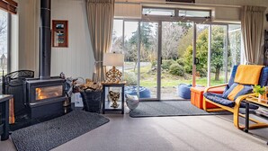 A cozy living room with a wood-burning stove, a comfortable sofa, and a view of the garden through large glass doors. - Alpine Way Cottage - Ohakune Holiday Home (Ohakune)