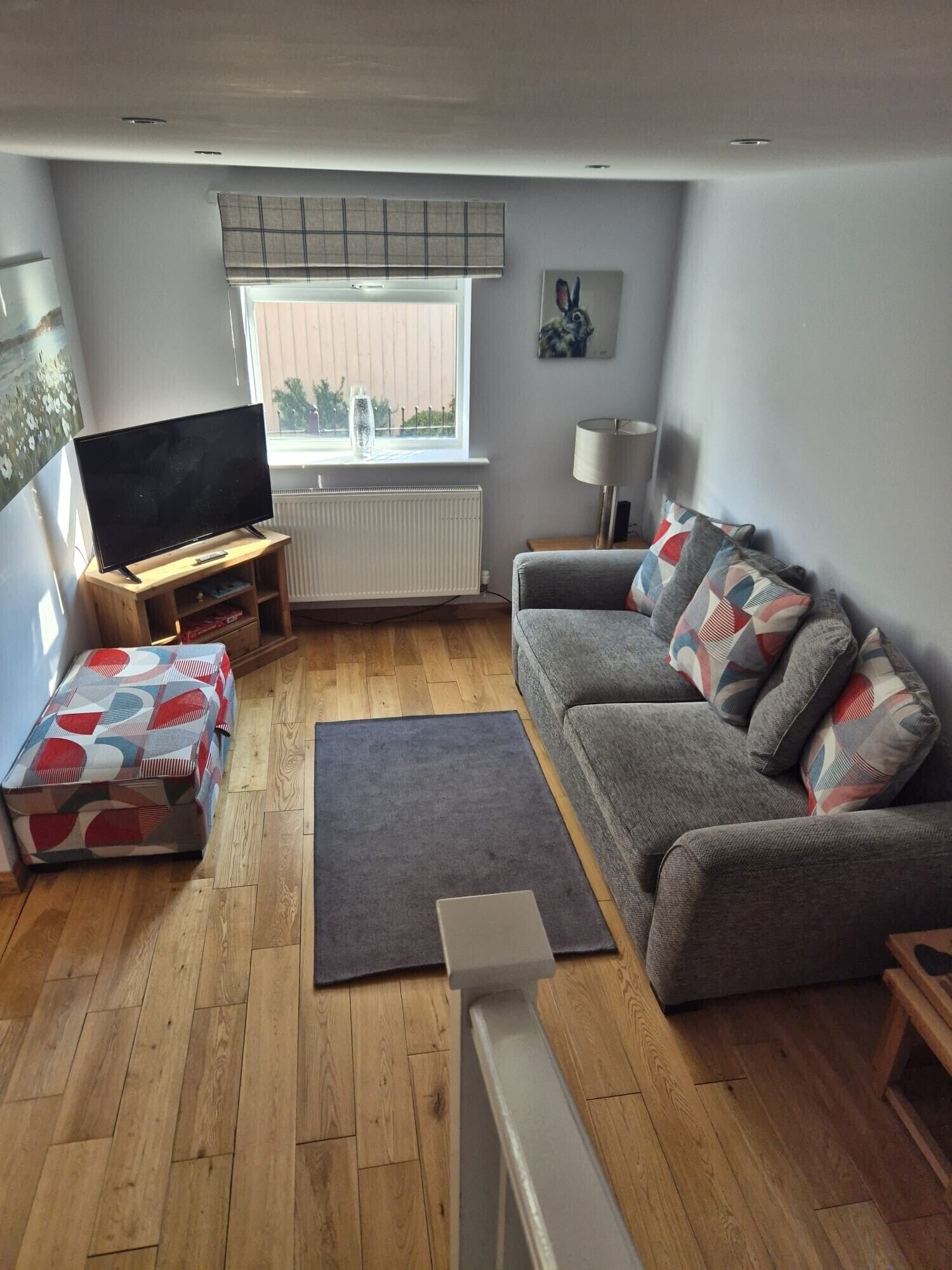 Una sala de estar con un sofá gris, un televisor de pantalla plana sobre un soporte de madera, una mesita de madera, una alfombra gris y una ventana con persianas.