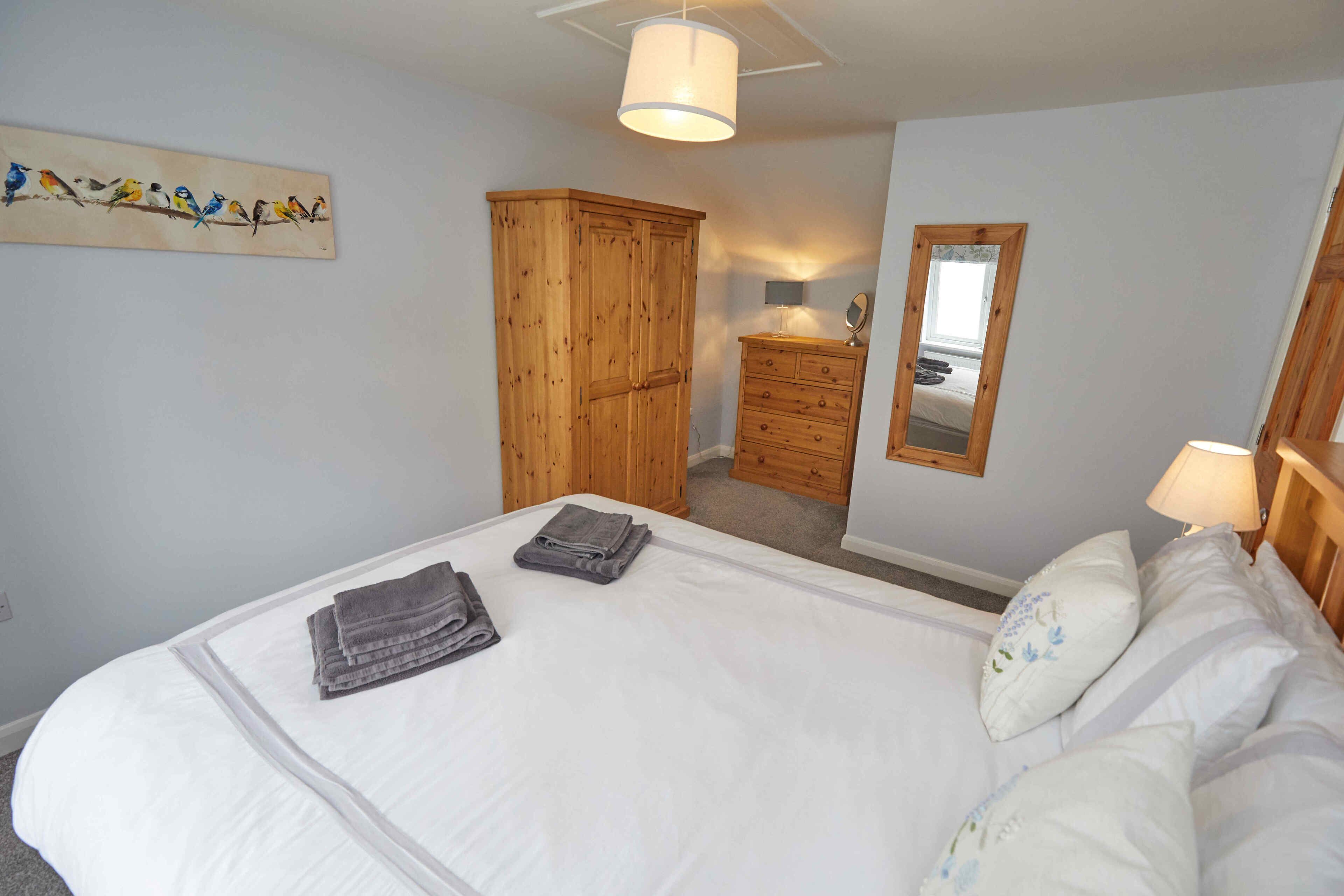 A neatly made bed with folded towels, a wooden wardrobe, a dresser, a mirror, and a wall-mounted artwork of birds.