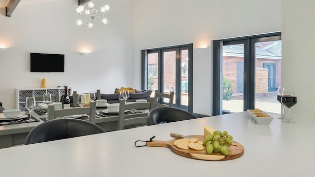 Un comedor moderno con una mesa grande dispuesta para una comida, un televisor montado en la pared y ventanas amplias que ofrecen vistas al exterior.