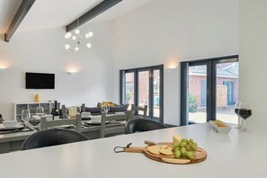 Uma sala de jantar moderna com uma mesa grande posta para refeição, uma televisão na parede e amplas janelas com vista para o exterior.