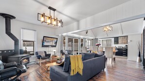 A spacious living area with a large gray sofa, a black leather chair, and a wooden floor. A black wood-burning stove is visible in the corner, and there is a dining area with a table and chairs. - Serene Lakefront Cottage Escape (Rivière-Rouge)