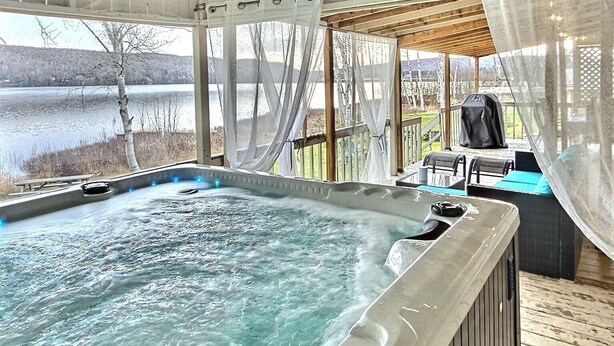 A hot tub in a covered outdoor area with a view of a lake and trees. - Serene Lakefront Cottage Escape (Rivière-Rouge)