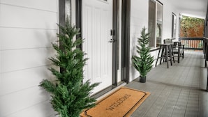 Un porche con un felpudo, dos árboles de Navidad en macetas y una zona de estar con sillas y una mesa.