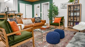 Una sala de estar acogedora con un sofá de cuero, sillones verde y naranja, una mesa de centro de vidrio y un taburete tejido azul.