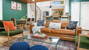 Una sala de estar con un sofá de cuero marrón, sillas verdes y anaranjadas, una mesa de centro de vidrio y techo de madera.