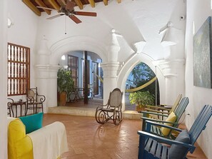 Ampla sala de estar com teto alto, vigas de madeira e ventoinha. Possui vários espaços de estar com cadeiras e um sofá. O cômodo apresenta paredes brancas, arcos de madeira e vista para uma área externa com plantas através de um arco.