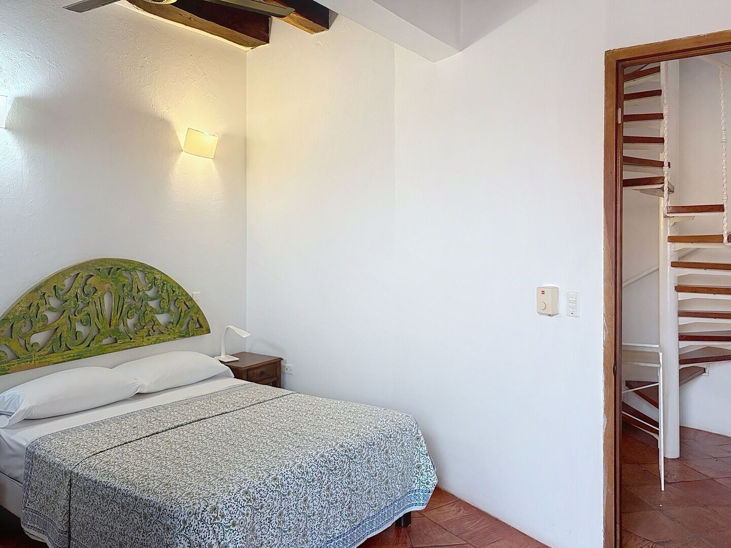 A bedroom with a bed, a nightstand, a wall-mounted lamp, and a staircase leading to an upper floor.