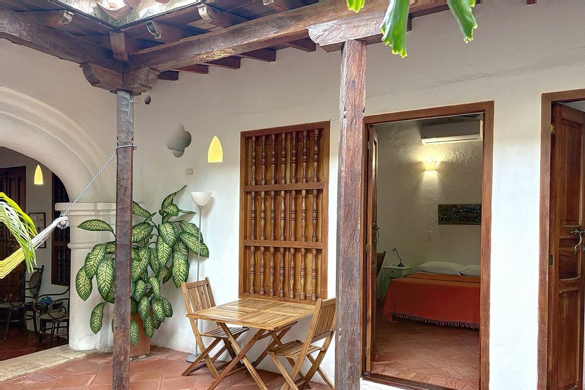 A wooden table and chairs in a covered outdoor area with a tiled floor, a large potted plant, and a wooden partition with a door leading to a room.