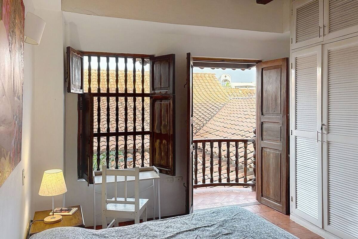 A bedroom with a bed, a desk, a chair, a lamp, and a view of the outside.
